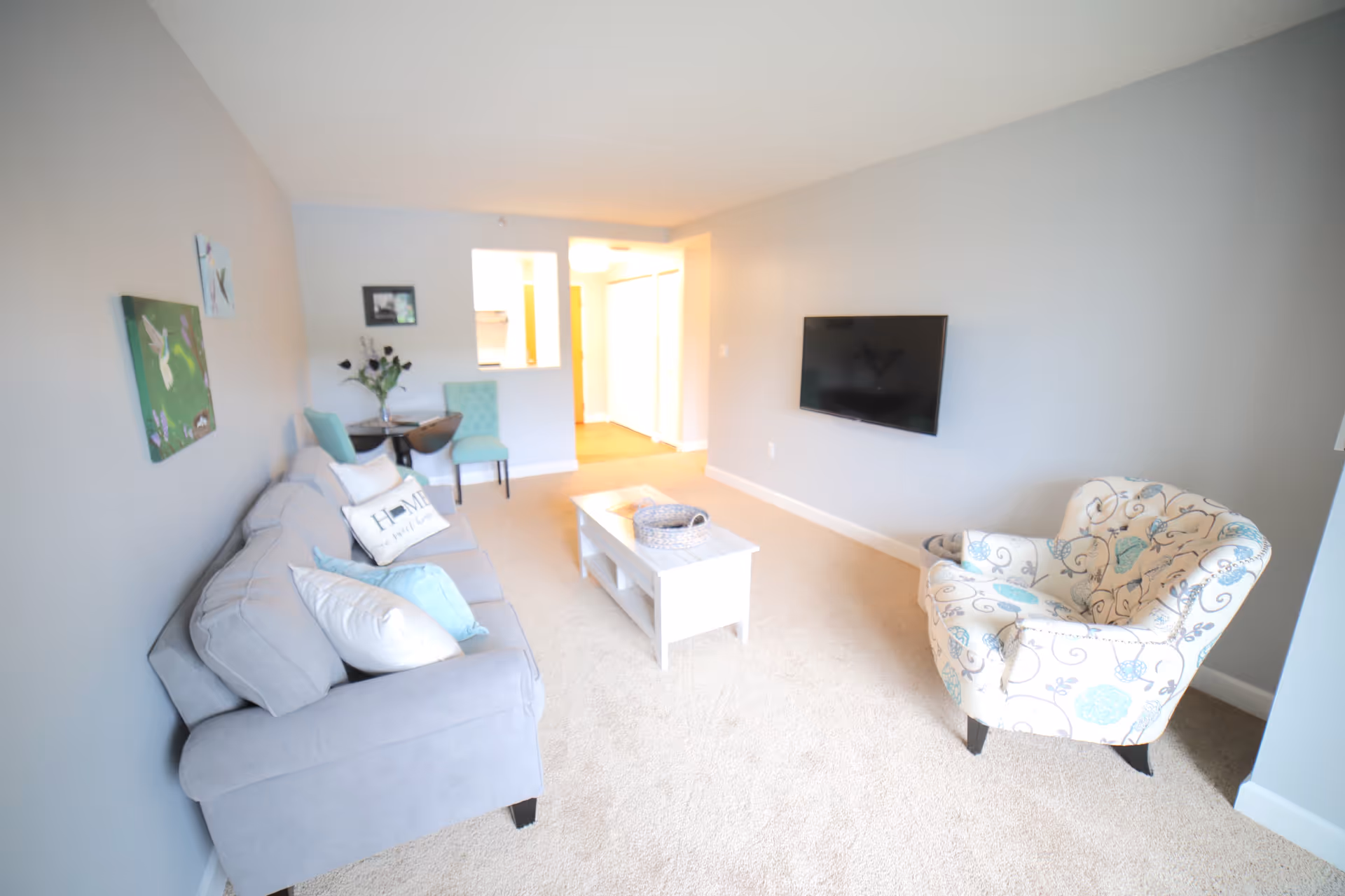 A bright living room with light beige carpet and gray walls. The room features a gray sofa with multiple pillows, a white coffee table with a decorative basket, and a patterned armchair with blue and beige floral designs. A flat-screen TV is mounted on the wall opposite the sofa. In the background, there is a small dining area with two turquoise chairs and a round table with a vase of flowers. A hallway with white closet doors is visible beyond the dining area.