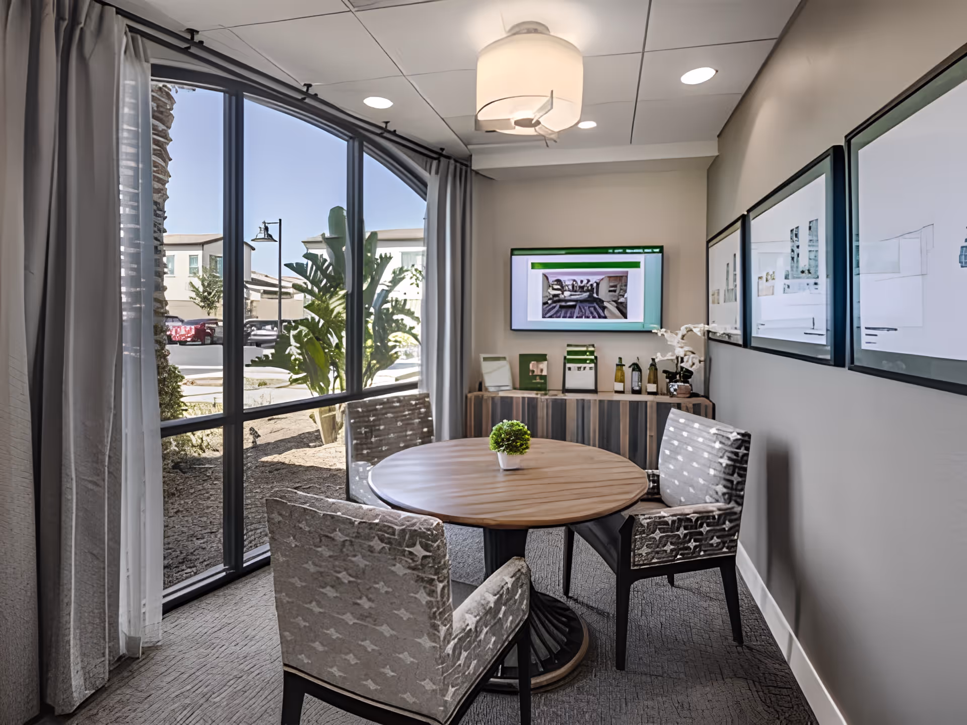 Small interior dining/meeting area with a round wooden table surrounded by upholstered chairs, a large arched window, wall-mounted TV and framed prints.