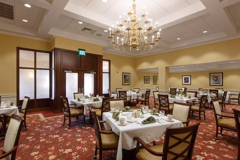 A formal dining room with multiple tables set for a meal, upholstered chairs, and a central chandelier.