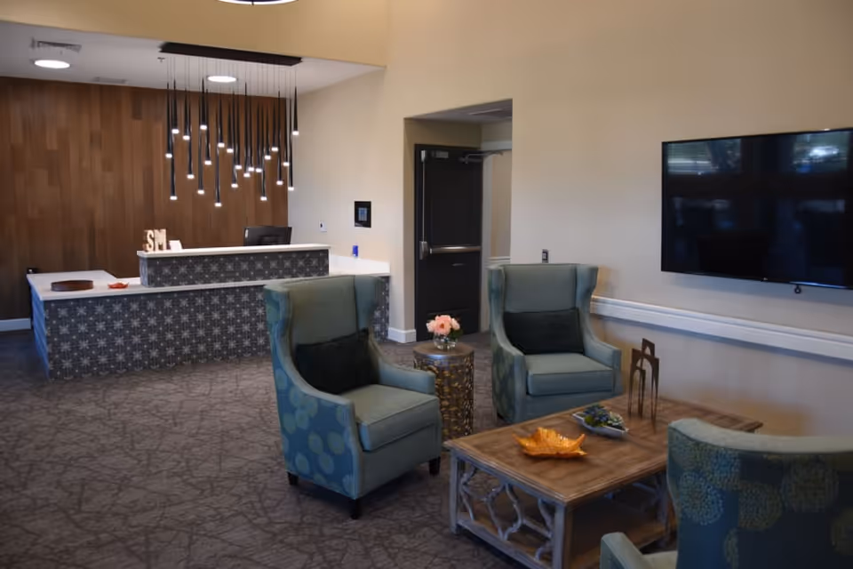 A modern reception area with a patterned front desk, hanging pendant lights, and a wood-paneled wall behind it. In the foreground, there are three upholstered armchairs with dark cushions arranged around a wooden coffee table decorated with a yellow leaf-shaped dish and other small decor items. A flat-screen TV is mounted on the wall to the right.