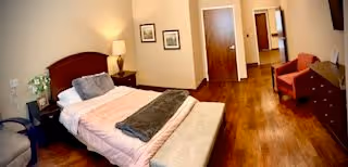 Bright bedroom with a single bed, bench, nightstand and lamp, dresser and chair on hardwood floors with framed pictures and an open doorway.