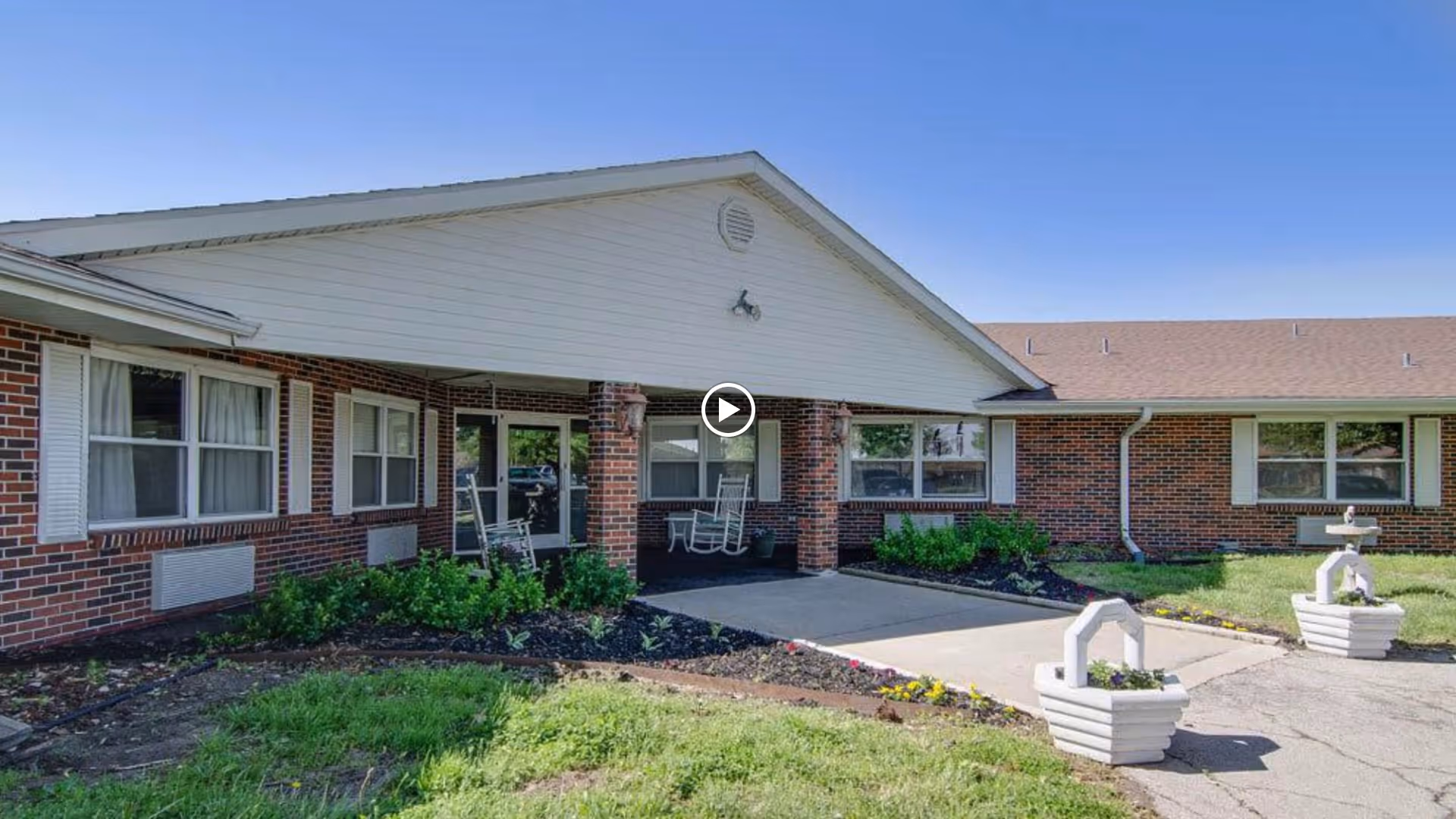 Exterior view of Osage Nursing Center showing a single-story brick building with white trim and a covered entrance. There are two white rocking chairs on the porch, flower beds with green plants and flowers, and a clear blue sky overhead.