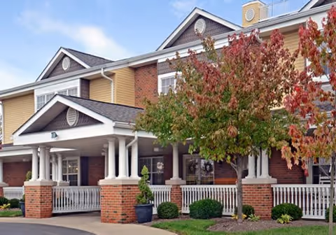 Front exterior view of a senior living facility building with a covered entrance supported by white columns, brick and beige siding, and a tree with green and red leaves in front.