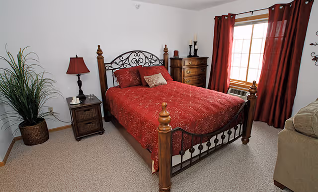 A bedroom with a wooden bed frame and red bedding, including pillows and a comforter. There is a wooden nightstand with a red lamp and a small decorative item on it. A wooden chest of drawers with candles and decorative items is placed against the wall near a window with red curtains. A green plant in a woven basket is in the corner, and part of a beige sofa is visible on the right side. The room has beige carpet and white walls.