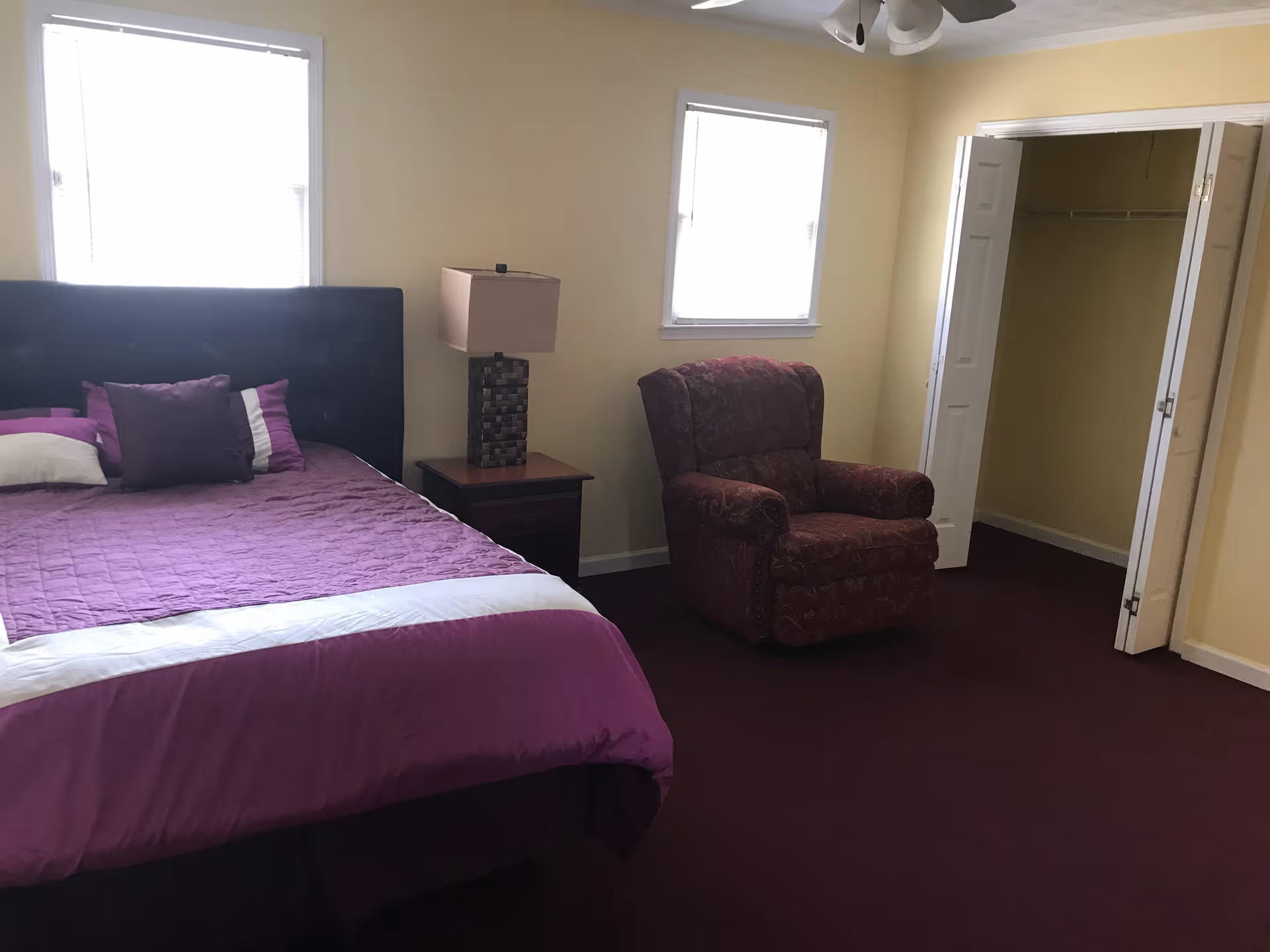 A bedroom with a large bed covered in purple bedding and pillows, a nightstand with a lamp, a patterned armchair, two windows with blinds, and an open closet with white bi-fold doors.