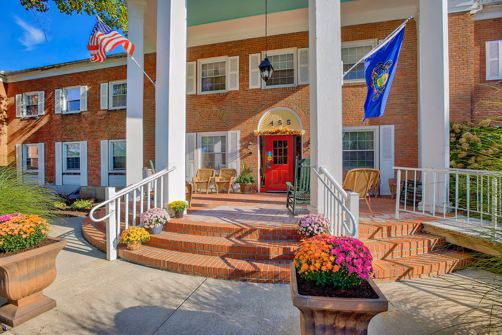 Front entrance of a brick building with white columns, a red door, and two flags on either side. There are steps leading up to the porch with potted flowers and outdoor chairs arranged near the door.