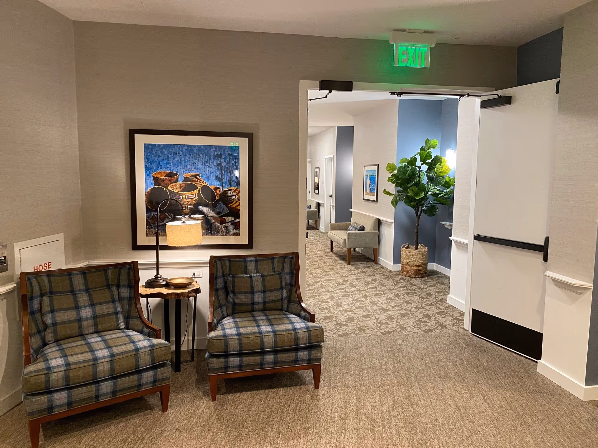 A cozy interior hallway area in a senior living facility with two plaid upholstered chairs and a small wooden side table with a lamp between them. Above the chairs is a framed artwork featuring pottery. The hallway extends into the background with additional seating, framed pictures on the walls, a large potted plant, and an open door with an illuminated green exit sign above it.