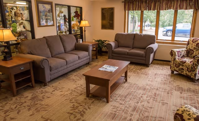 A cozy living room with two gray sofas, a floral patterned armchair, a wooden coffee table with a newspaper on it, two side tables with lamps, large windows with a view of parked cars outside, and framed pictures on the walls.