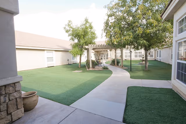 Outdoor courtyard area at Brookdale North Gilbert with a concrete pathway, artificial grass, several trees, a pergola with a swing, and surrounding building walls with windows.