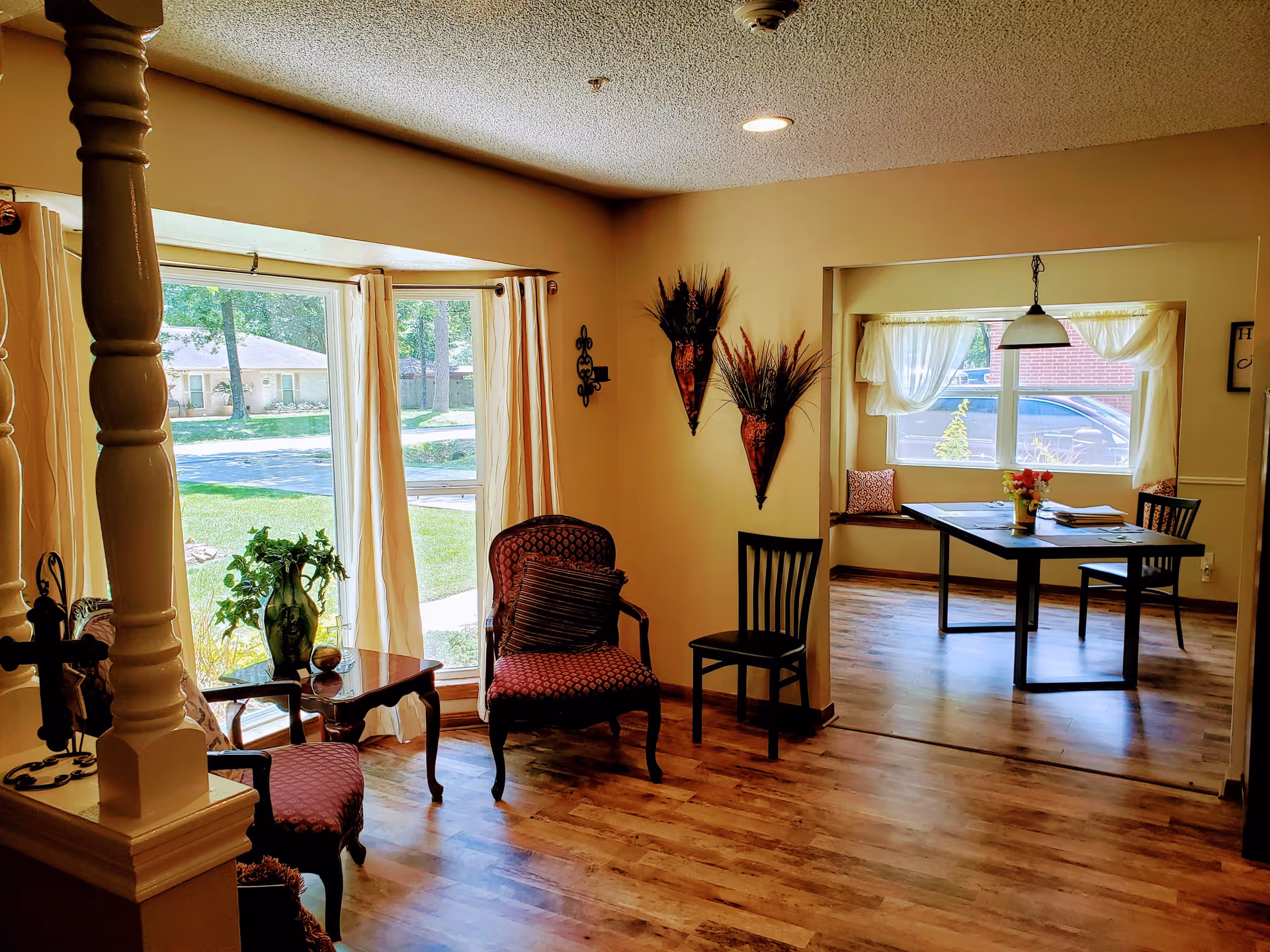 A cozy interior space with wooden flooring featuring a sitting area with two upholstered chairs and a small table with a green vase and plant near a large window with cream curtains. Adjacent to this is a dining area with a rectangular table, two chairs, a window seat with cushions, and a hanging light fixture above the table. The walls are decorated with two wall-mounted vases containing dried plants.