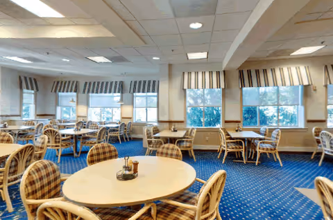 A bright dining room with multiple round tables and cushioned chairs arranged neatly. The room has large windows with striped valances allowing natural light to fill the space. The floor is carpeted in blue with a subtle pattern, and the ceiling has recessed lighting.