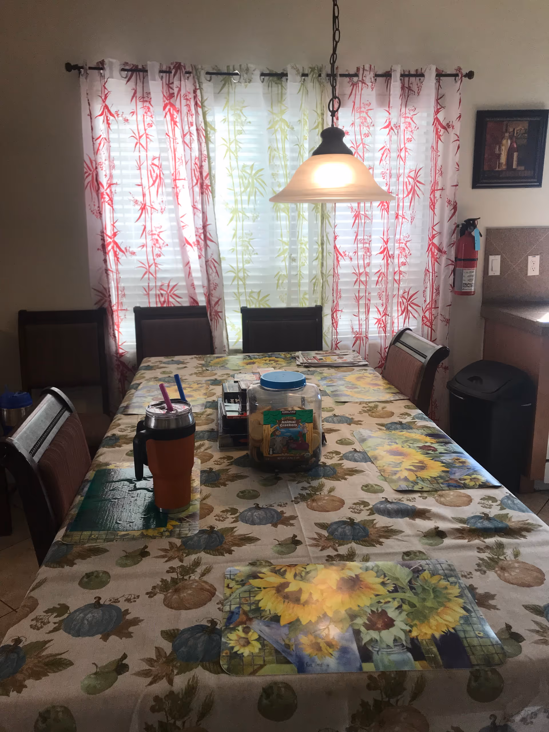 Dining area with a rectangular table covered with a floral and pumpkin-themed tablecloth. The table has placemats featuring sunflowers, a container of animal crackers, a couple of mugs with straws, and a small organizer. Behind the table are chairs and a window with white blinds and red and green patterned curtains. A hanging light fixture is above the table. On the right wall, there is a fire extinguisher, a framed picture, and part of a kitchen counter with a trash bin underneath.