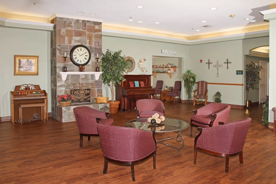 A cozy living room area with four maroon upholstered chairs arranged around a glass coffee table. The room features a stone fireplace with a large clock above it, a wooden piano, and several potted plants. The walls are light green with decorative crosses and framed artwork. The floor is wooden, and the ceiling has recessed lighting.
