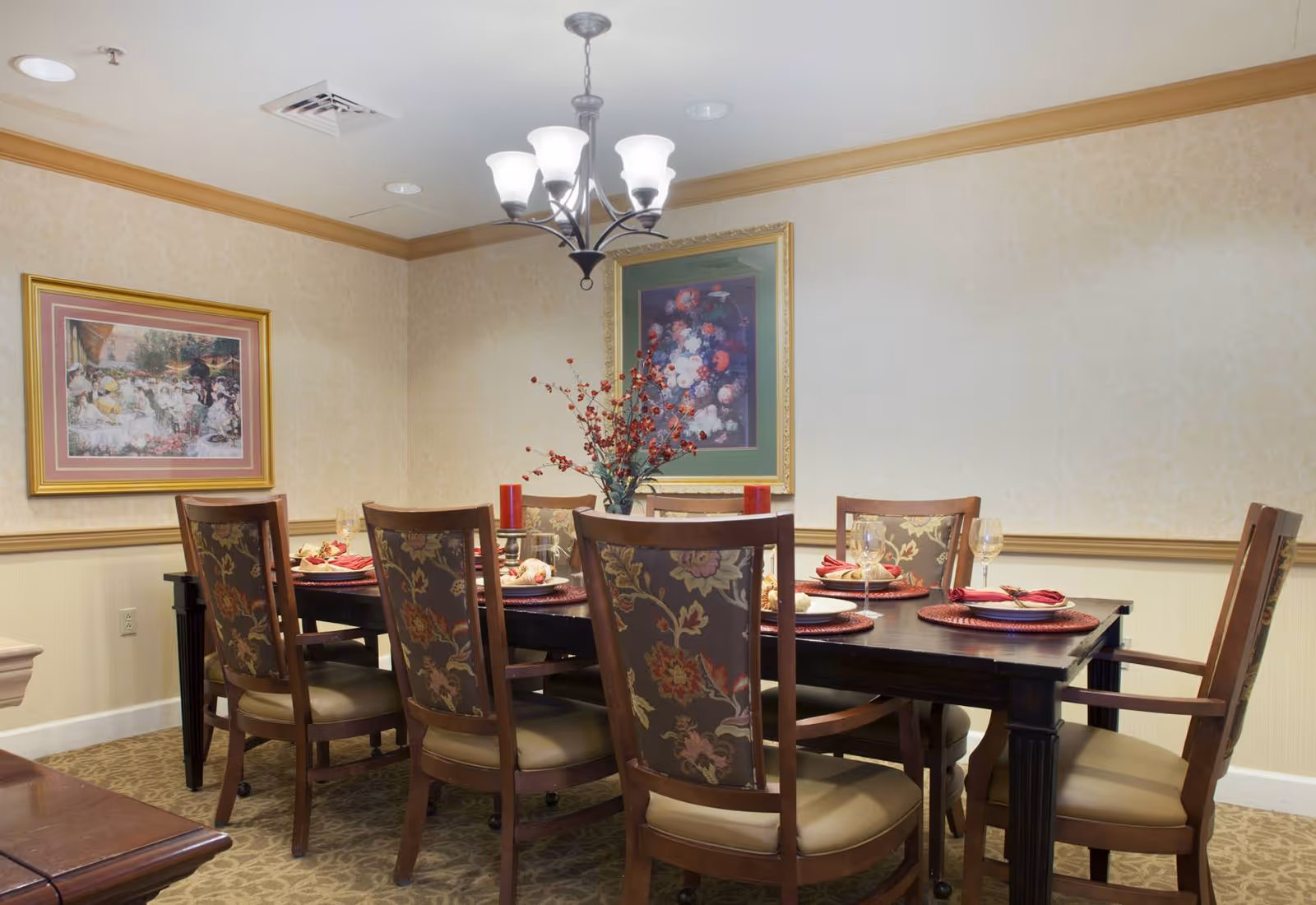 A formal dining room with a dark wood table set for six, patterned chairs, place settings, a floral centerpiece and a hanging chandelier.