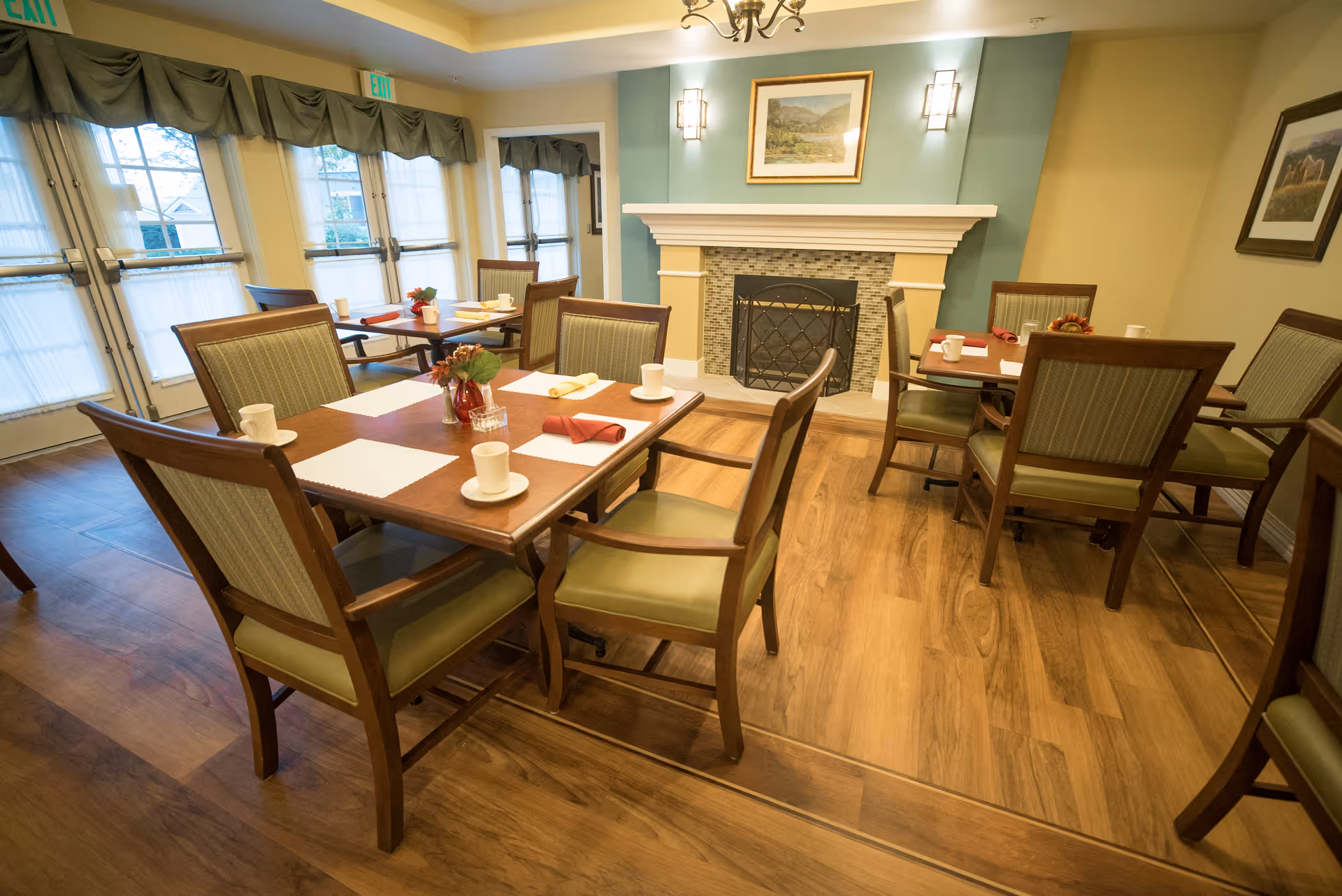 A bright dining room with wooden tables and chairs set with cups and napkins in front of a fireplace.