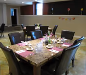A dining area with a square marble table set for six people with white cups, saucers, and pink placemats. The chairs are dark brown leather, and there are small flower arrangements on the table. The background shows a partial wall with colorful flower decorations and a window letting in natural light.