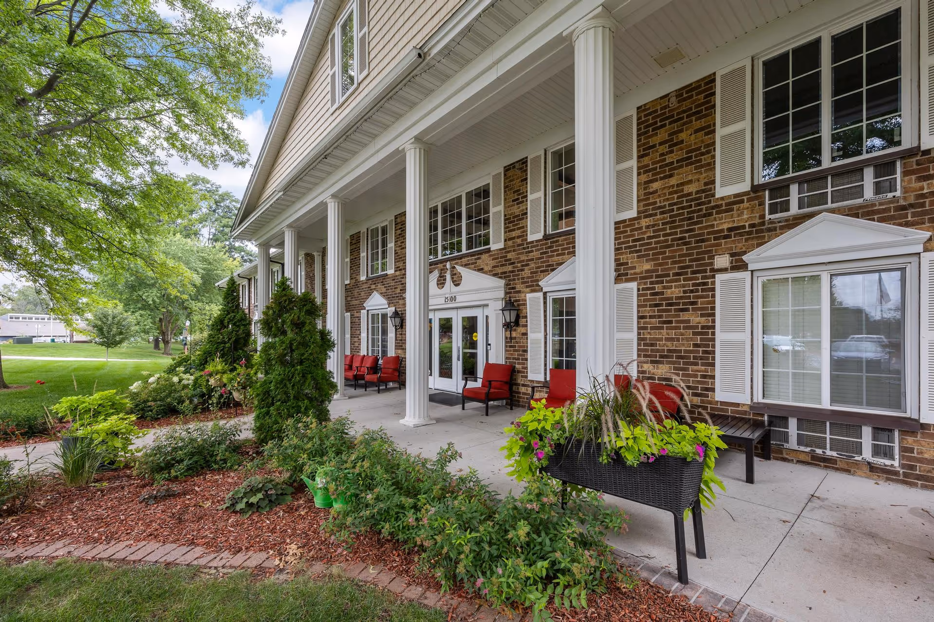 Front entrance of a brick senior living building with white columns, a covered porch with red chairs, and landscaped greenery.