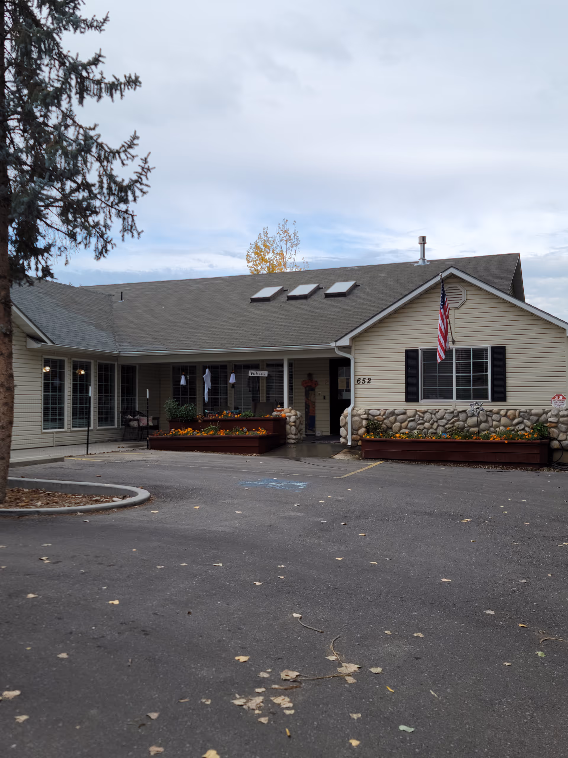 Exterior view of a single-story building with beige siding and a stone facade at the lower part. The building has a gray roof with three skylights and an American flag mounted near a window. There are flower beds with colorful flowers in front of the entrance, and a paved parking area with some fallen leaves. A tree is visible on the left side of the image.