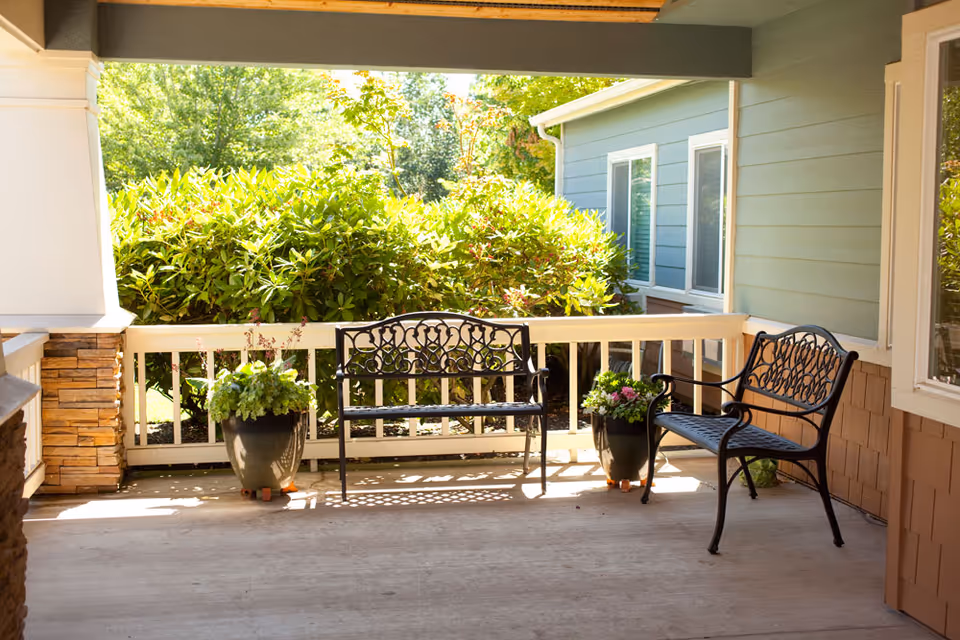 Covered outdoor patio area with two black metal benches and two potted plants, surrounded by greenery and adjacent to a building with blue siding and white trim.