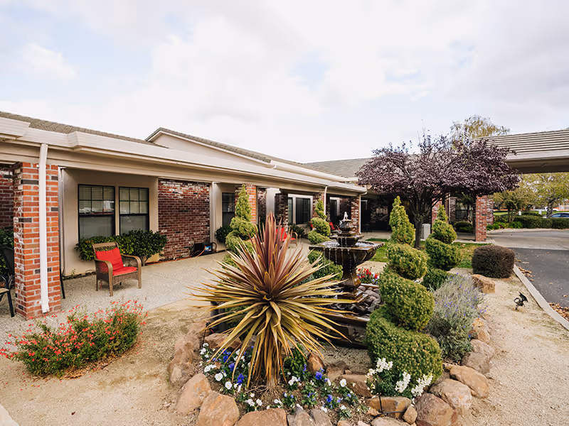 Outdoor view of a senior living facility with a landscaped garden featuring a multi-tiered water fountain, various shrubs, and flowering plants. The building has brick walls and large windows with a covered walkway. There are chairs placed near the building entrance and a driveway visible on the right side.