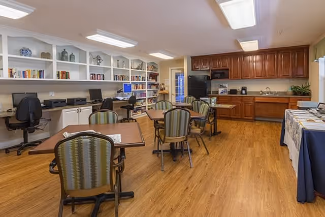 A spacious room with wooden flooring featuring several tables and chairs arranged for seating. On the left side, there are built-in shelves filled with books and decorative items, along with a desk area equipped with computers and office chairs. The back wall has a kitchen area with wooden cabinets, a black refrigerator, a microwave, a coffee maker, and a sink. A table covered with a cloth is positioned on the right side of the room.