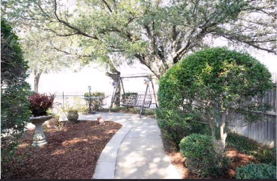A serene outdoor garden path with a curved concrete walkway surrounded by neatly trimmed bushes, a birdbath with plants, and a large tree providing shade. A wooden swing bench is visible in the background near a fence.