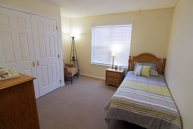 Small bedroom with a single bed, wooden headboard, nightstand, dresser, closet, armchair, and a window with blinds.