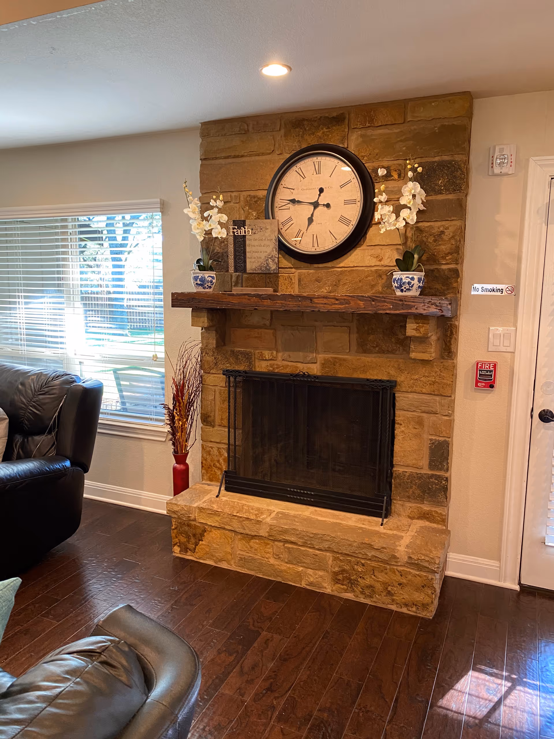 A cozy living room area featuring a stone fireplace with a wooden mantel. On the mantel, there is a large round clock with Roman numerals, two potted white orchid plants in blue and white pots, and a decorative plaque with the word 'Faith'. To the left, there is a window with blinds partially open, letting in natural light. A black leather armchair is partially visible in the foreground. The floor is dark hardwood, and there is a 'No Smoking' sign and a fire alarm on the wall to the right of the fireplace.