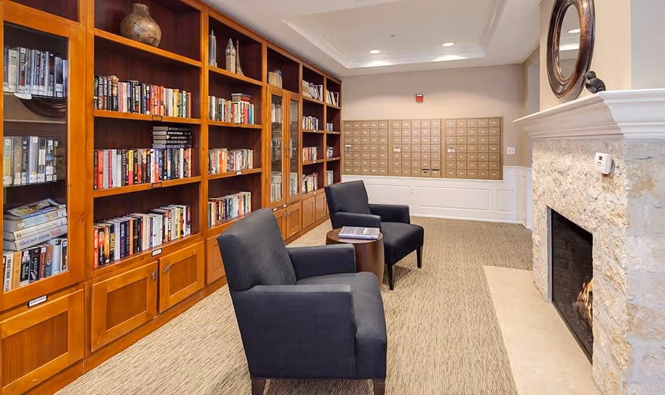 A cozy library room with wooden bookshelves filled with books along the left wall, two dark upholstered armchairs with a small round table between them, and a stone fireplace on the right side. In the background, there are multiple mailboxes mounted on the wall.