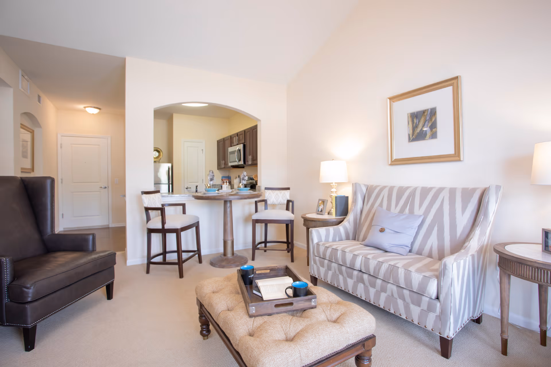 A bright and cozy living room area with a patterned sofa, a cushioned ottoman with a tray holding a book and two blue mugs, a dark leather armchair, and two side tables with lamps. In the background, there is a small round dining table with two chairs and an open view into a kitchen with dark cabinets and stainless steel appliances.