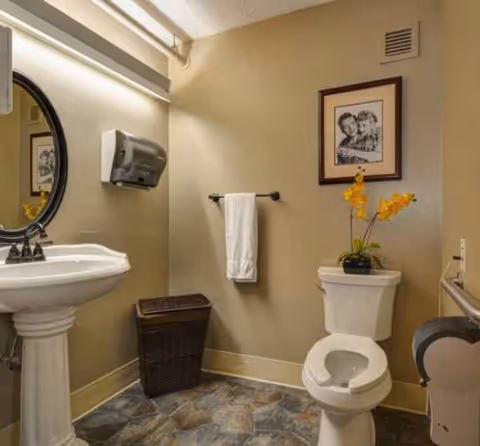 A clean bathroom with pedestal sink, mirror, toilet, towel, framed art, and a potted yellow flower.