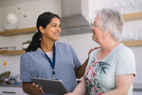 A smiling caregiver in blue scrubs holding a tablet and gently touching the shoulder of an elderly woman with short white hair and glasses, standing together in a bright kitchen with white tiled walls and wooden shelves.