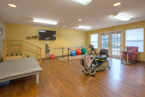 A rehabilitation room with wooden flooring and yellow walls featuring exercise equipment including a recumbent bike, parallel bars, therapy balls, a padded bench, and a small staircase with handrails. There is a wall-mounted TV and double glass doors letting in natural light.