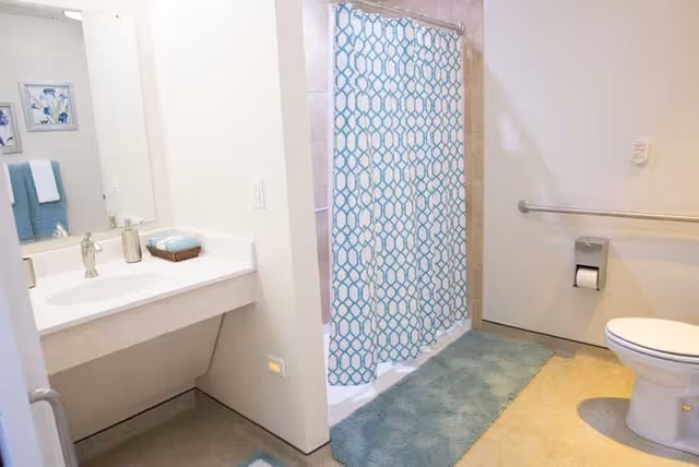A clean and well-lit bathroom featuring a white sink with a silver faucet and soap dispenser, a mirror above the sink, a shower area with a blue and white patterned curtain, a toilet with a grab bar on the wall, and blue and yellow bath mats on the floor.