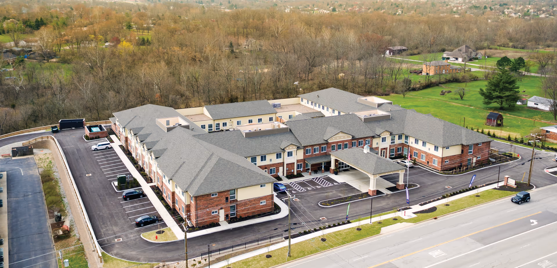 Aerial view of a large two-story brick and beige senior living facility with a covered front entrance, parking lot, and surrounding roads and trees.