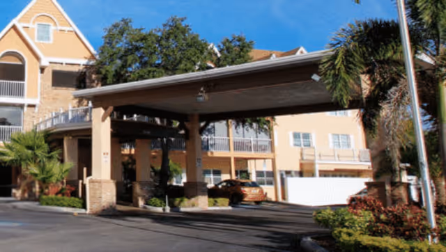 Exterior view of Bayside Terrace senior living facility showing a covered entrance with columns, a driveway, parked cars, and surrounding palm trees and landscaping under a clear blue sky.