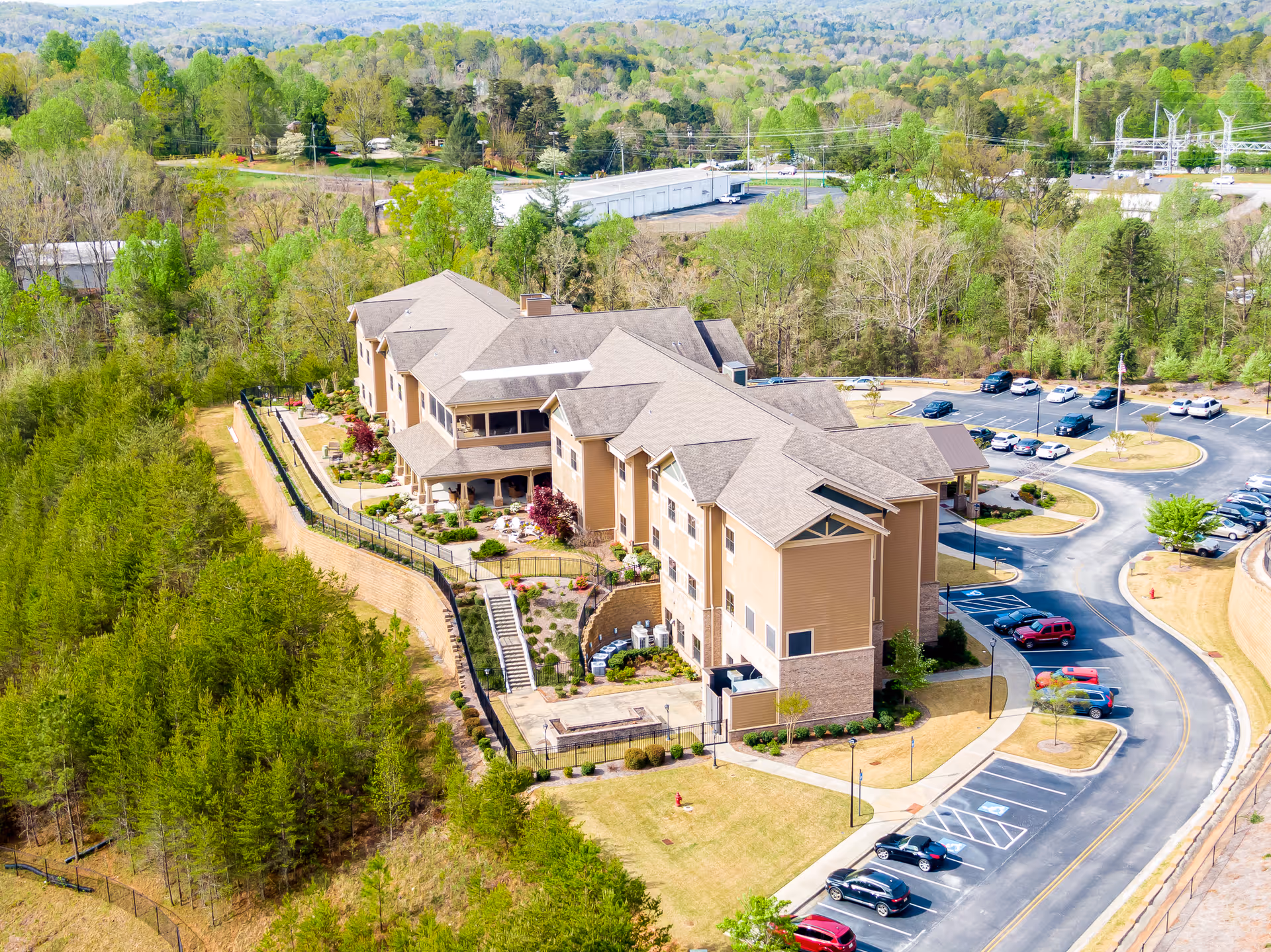 Aerial view of Chelsey Park Health & Rehabilitation building surrounded by trees and greenery. The multi-story facility has a beige exterior with a parking lot filled with cars nearby. The landscape includes well-maintained lawns, pathways, and a fenced garden area.