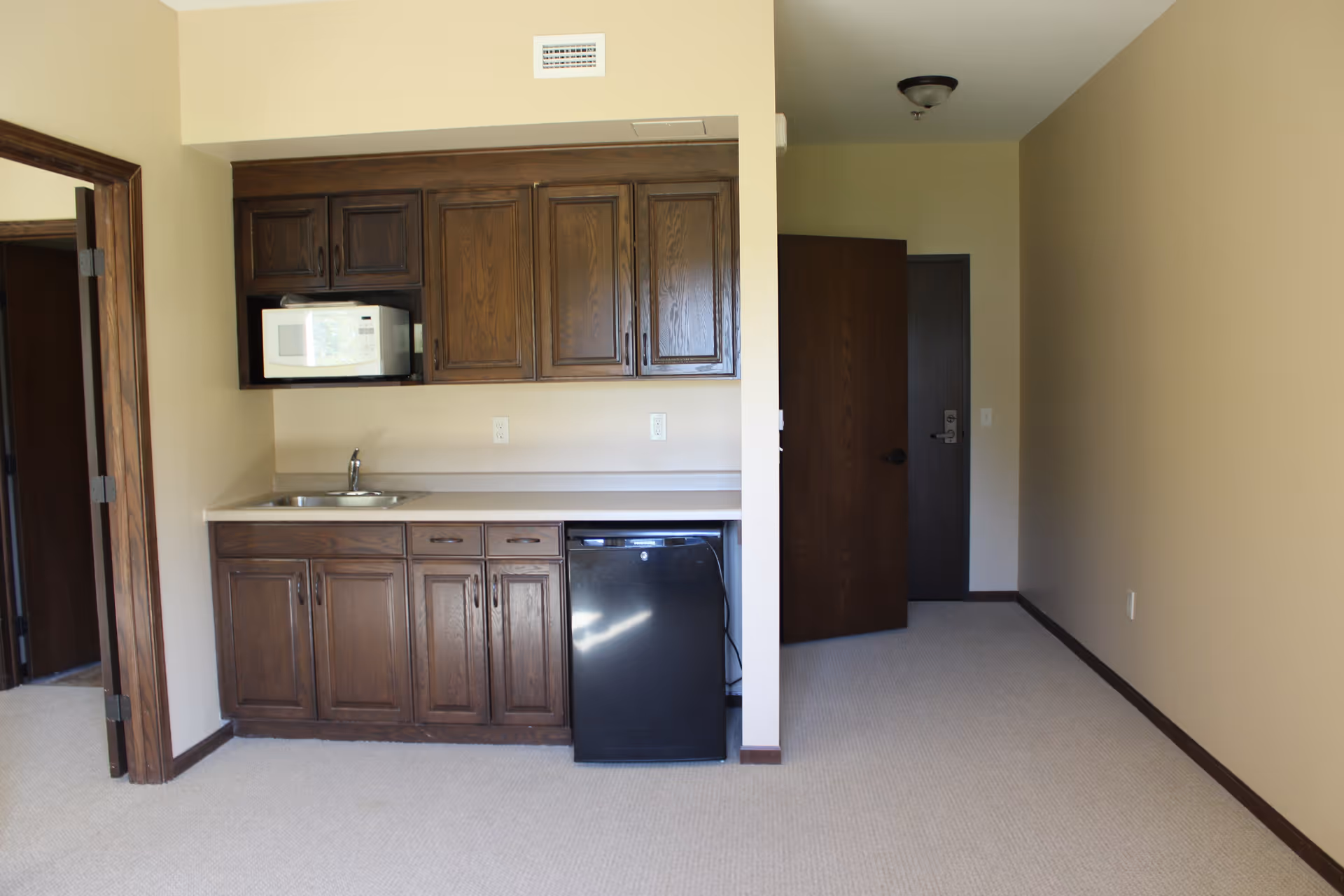 Interior view of a small kitchenette area with dark wooden cabinets, a white microwave, a small sink, and a black mini refrigerator. The room has beige walls and carpeted floor, with an open doorway leading to another room and a closed door in the background.