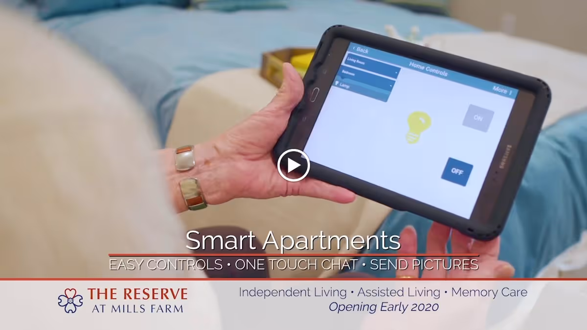 A person holding a tablet device displaying home controls for a smart apartment, with options to turn lights on or off. The background shows a bed with blue bedding.