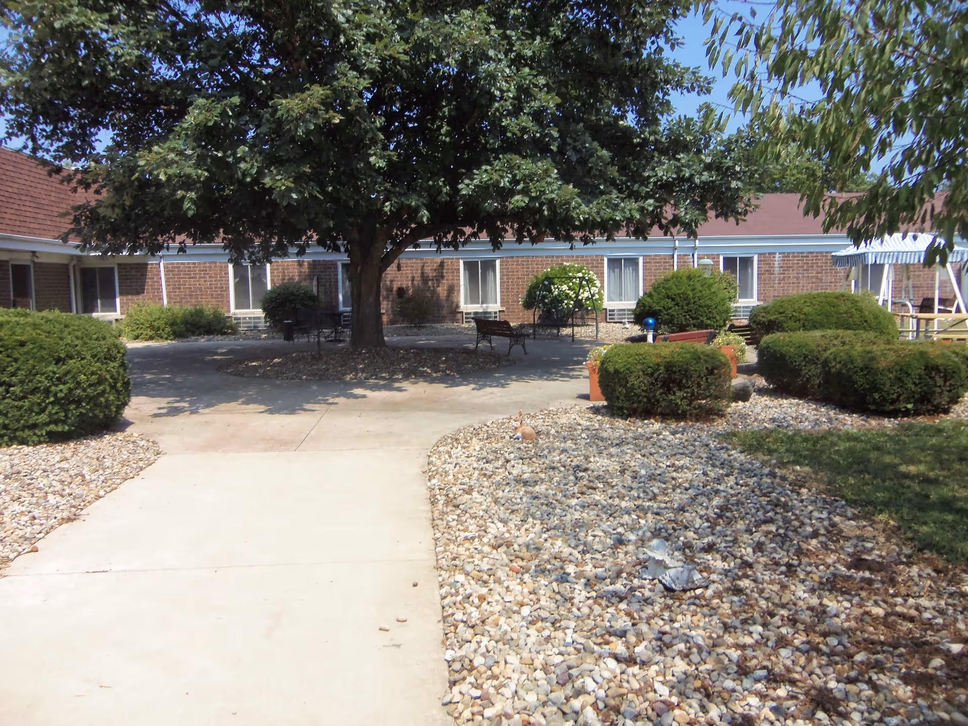 Outdoor courtyard area at a senior living facility with a large tree providing shade over benches and tables. The courtyard is surrounded by a single-story brick building with windows and a red roof. There are neatly trimmed bushes and a rock garden lining the concrete walkway leading into the courtyard.