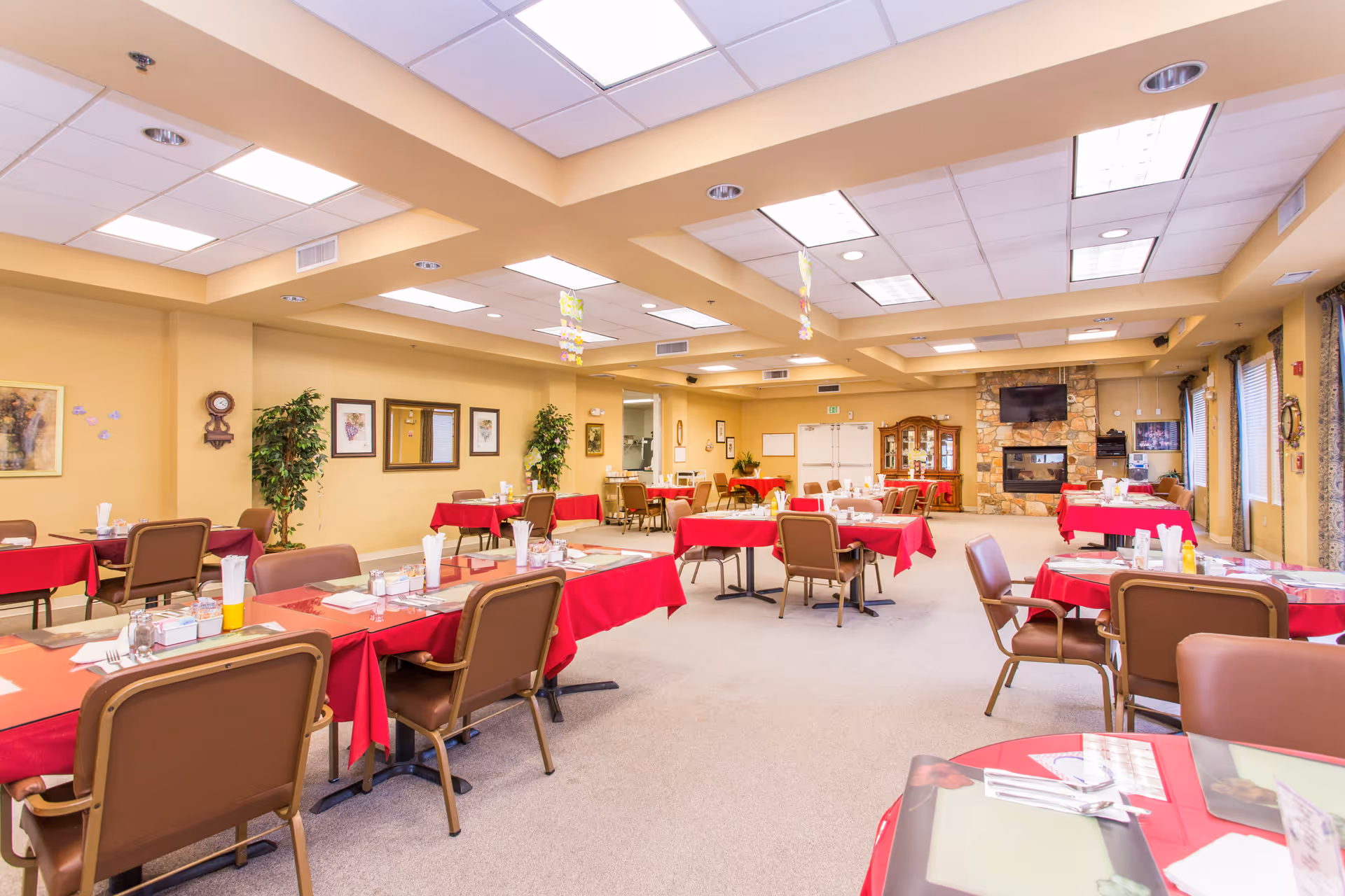 A spacious dining room with multiple tables covered in red tablecloths, each set with placemats, napkins, utensils, and condiments. Brown cushioned chairs surround the tables. The room has beige walls adorned with framed pictures and a clock, potted plants, and a stone fireplace with a mounted TV above it. Large windows with curtains allow natural light to enter the room.