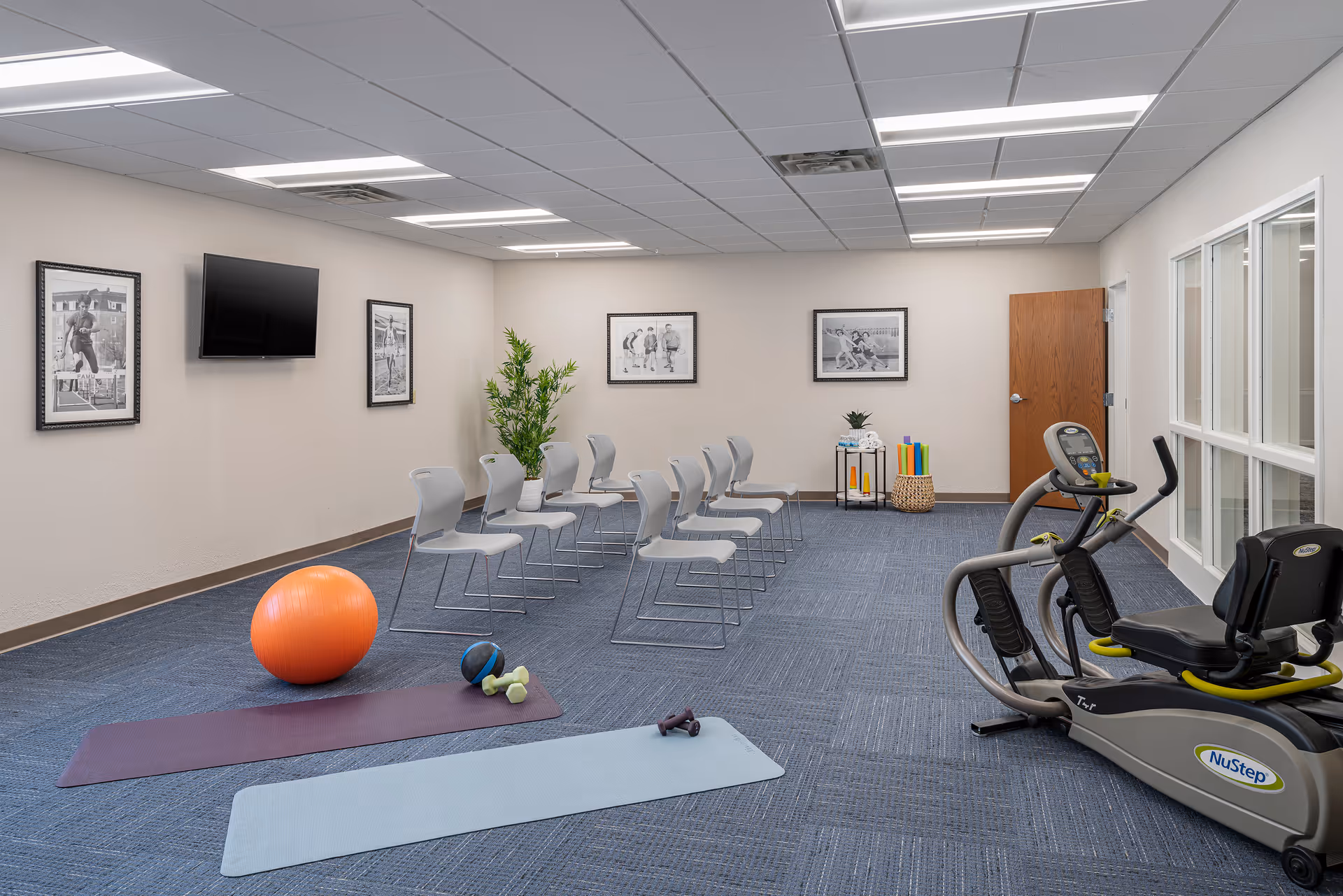 A fitness and activity room with exercise equipment including a NuStep machine, two yoga mats, an orange exercise ball, small dumbbells, and a medicine ball. There are two rows of gray chairs arranged in the middle of the room, framed black and white sports photos on the walls, a flat screen TV mounted on the wall, a potted plant, and a small table with colorful foam rollers near a wooden door.
