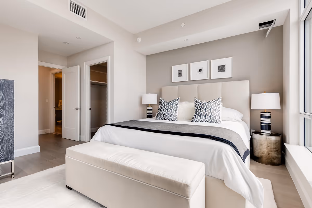 A modern bedroom with a large bed featuring a padded headboard, white bedding, and decorative pillows. There are two bedside tables with matching lamps, three framed artworks above the bed, a cushioned bench at the foot of the bed, and a large window letting in natural light. An open door reveals a walk-in closet and a glimpse of a bathroom.