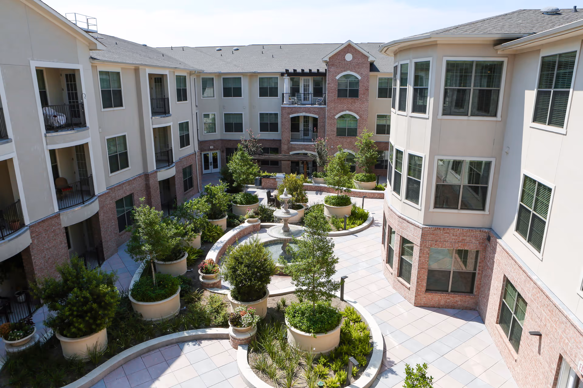 View of a courtyard garden area within a senior living facility, featuring large potted plants, a central water fountain, and surrounding multi-story buildings with balconies and windows.
