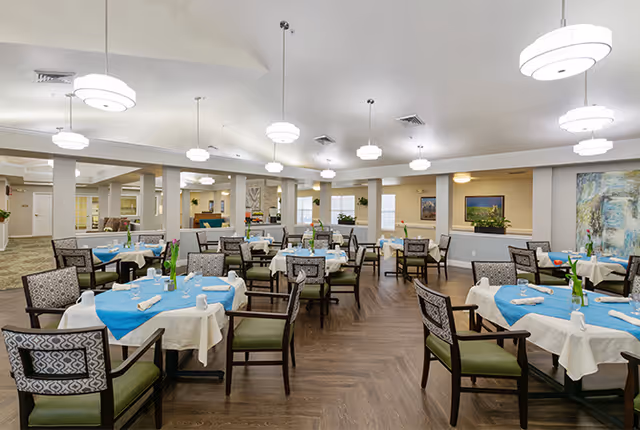 A spacious dining room in a senior living facility with multiple tables covered in white and blue tablecloths, each set with cups, plates, and napkins. The room features wooden flooring, cushioned chairs with patterned backs, modern pendant lights hanging from the ceiling, and artwork on the walls.