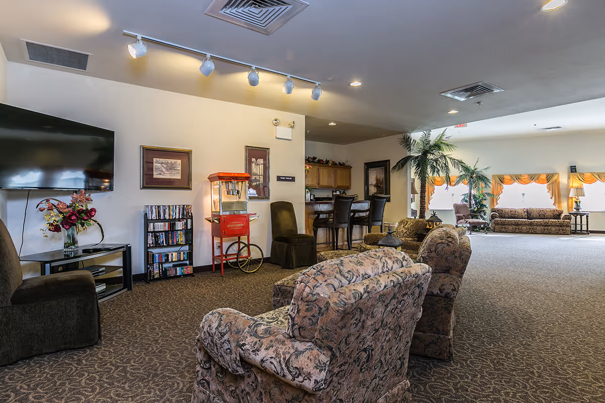 Spacious senior living common area with patterned armchairs and sofas, a wall-mounted TV, popcorn machine, bookcase, and bar seating.