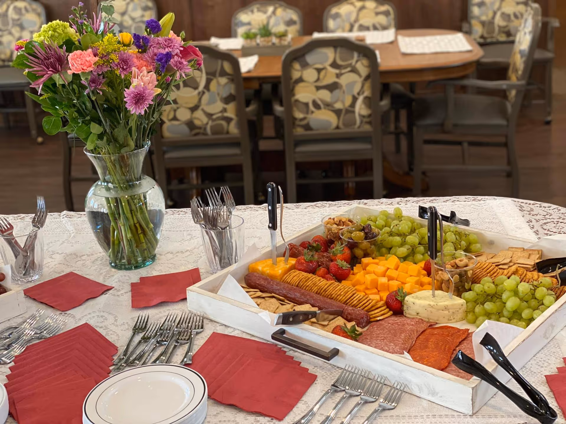 A table set with a large tray of assorted snacks including grapes, strawberries, cheese cubes, crackers, and sliced meats. There are utensils in clear cups, red napkins, and a vase with a colorful bouquet of flowers on the table. In the background, there are patterned chairs and a wooden dining table.