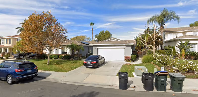 A suburban street view showing a single-story house with a two-car garage and a driveway with a black car parked on it. The house is surrounded by a green lawn, trees, and shrubs. Several trash bins are lined up on the curb in front of the house. Neighboring houses and cars are visible on either side under a clear blue sky.