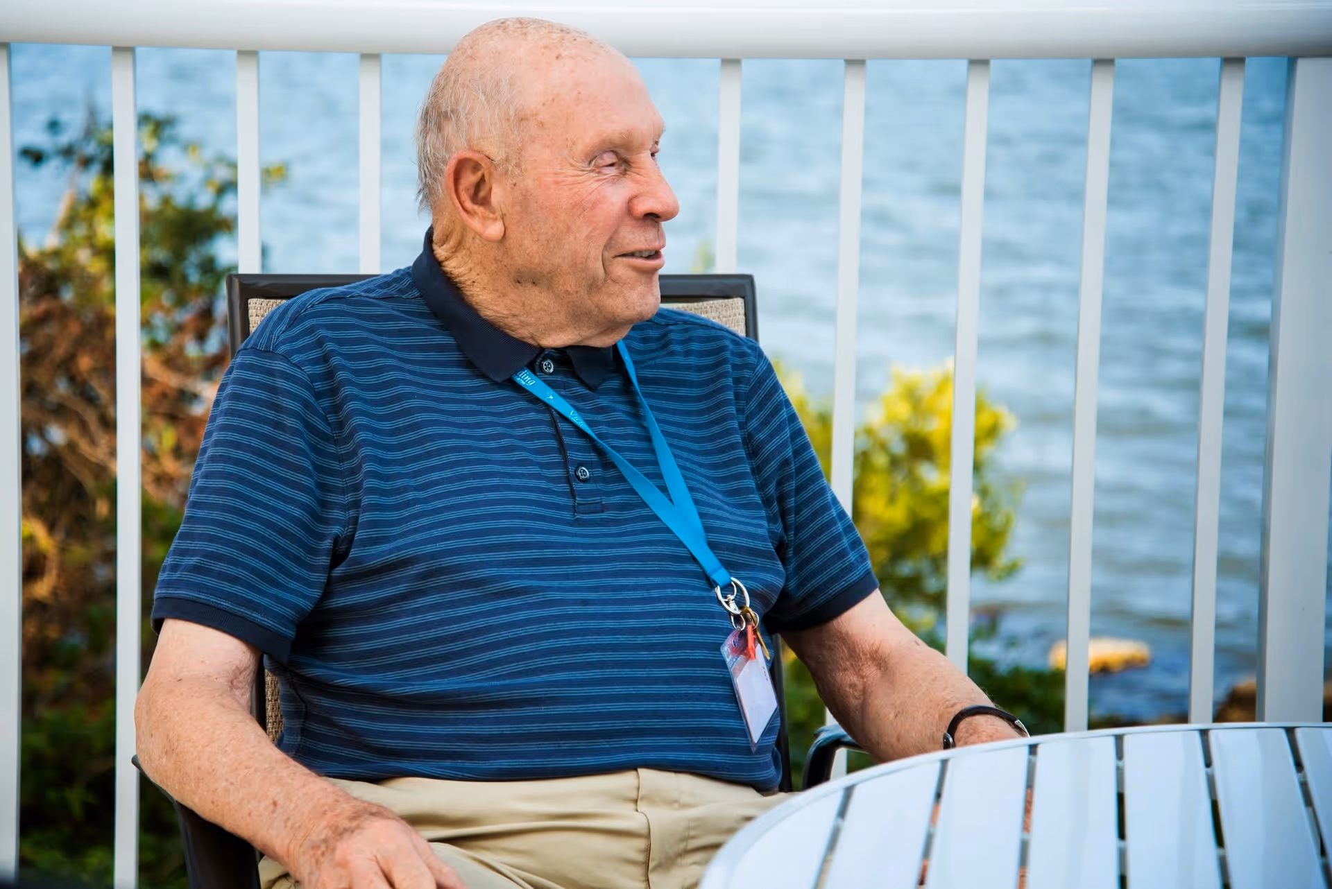 An elderly man wearing a blue striped polo shirt and beige pants sits on a chair outdoors near a white railing, with a body of water and greenery in the background. He has a blue lanyard with an ID badge around his neck and is looking to his left.