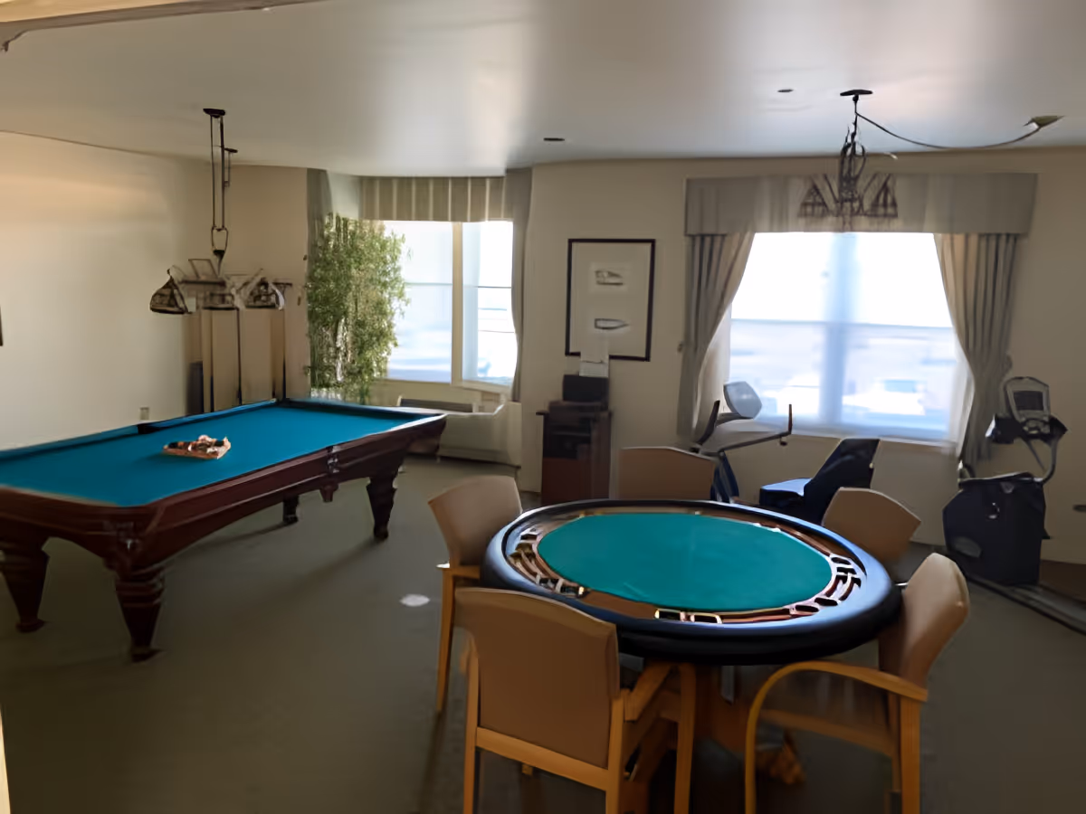 A recreational room with a pool table, a round card table with chairs, two exercise bikes near a large window with curtains, and a potted plant in the corner.